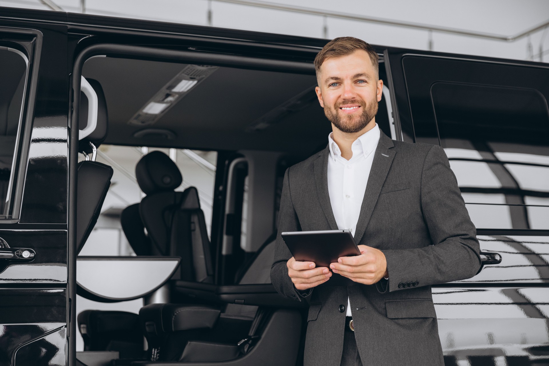 Smiling friendly car seller in suit standing in car salon and holding tablet. It's always pleasure to buy a car on a right place.